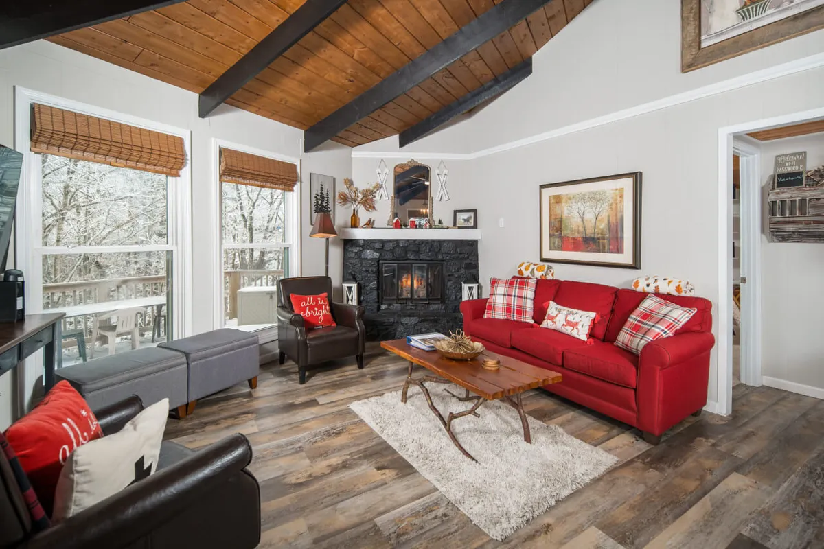 A warm living space with a red sofa, black fireplace, wooden beams, and large windows letting in natural light. The view through the windows suggests a close connection to the outdoor environment.