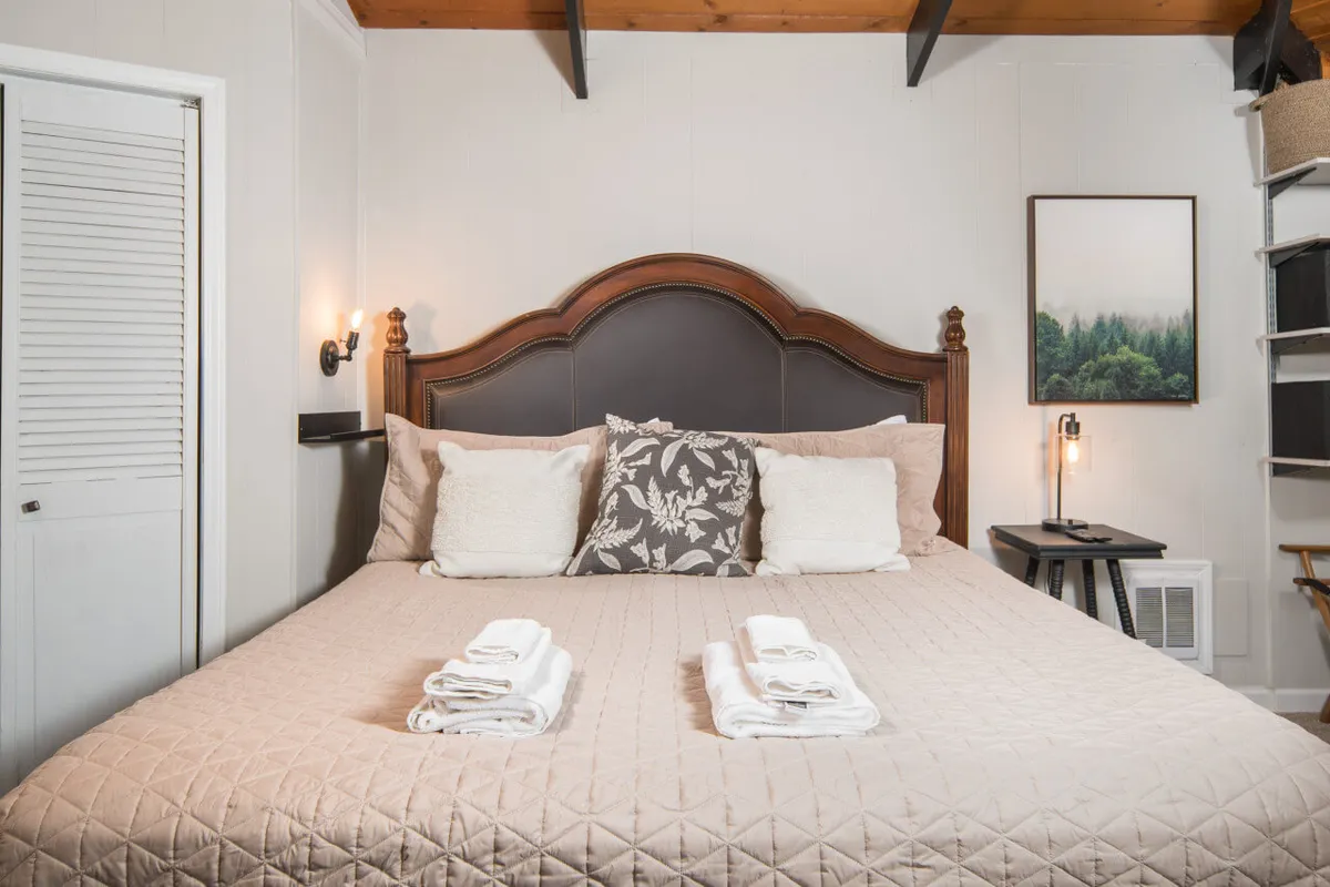 This image shows a serene bedroom with a bed featuring an elegant headboard, decorative pillows, nightstand with a lamp, and calming nature-themed artwork on the wall. Natural light bathes the room, enhancing the tranquil and restful atmosphere.