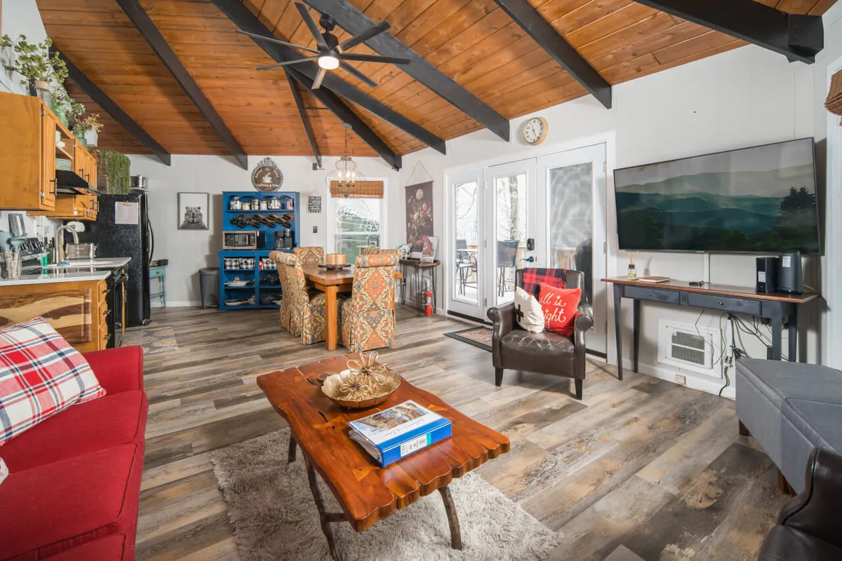 A cozy, open-concept living area featuring a red couch, wooden dining table, blue bookshelf, and, most importantly, a large window that frames an outdoor view—likely to the surrounding mountains or natural landscape. The room glows with natural light and feels connected to the outdoors, inviting relaxation after a day of adventure.