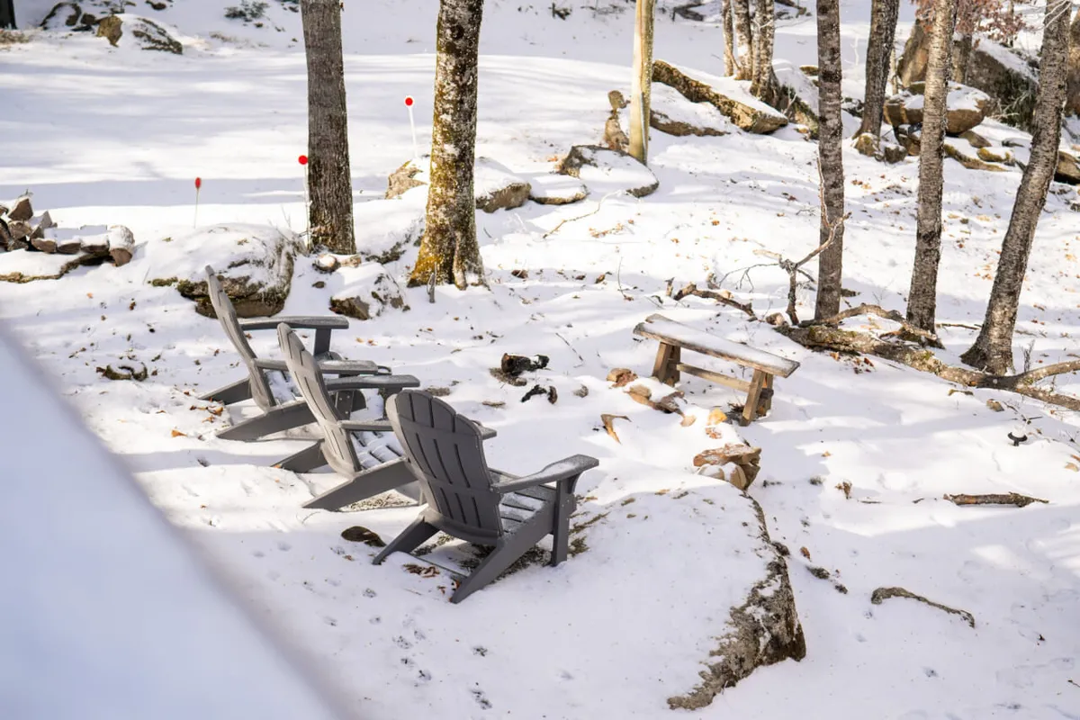 An outdoor seating area with wooden chairs and a small table on a snow-covered ground, surrounded by trees. The setting is peaceful, perfect for enjoying nature or relaxing with a pet at your feet.