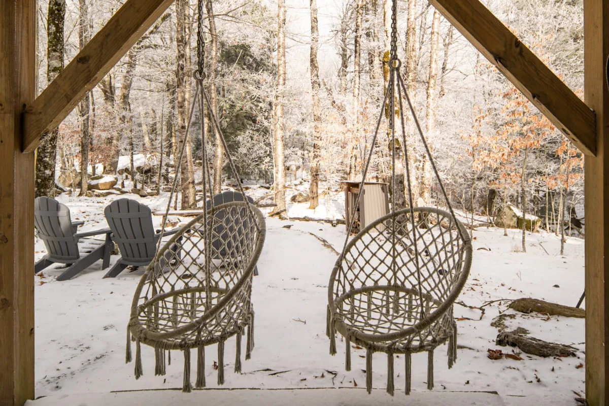 The photo features hanging chairs overlooking a snowy landscape, with forested surroundings creating a serene and inviting outdoor retreat. The placement suggests a perfect spot for reading, relaxing, or gathering, and hints at an inviting outdoor fire pit nearby.