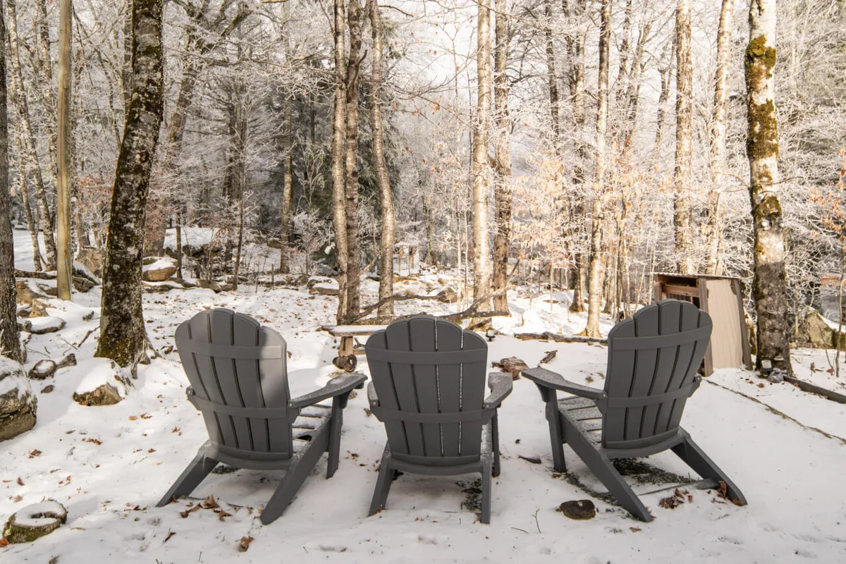 A cluster of adirondack chairs sits on a snow-covered ground surrounded by trees, creating an intimate setting for gathering outdoors, enjoying drinks, or stargazing under clear mountain skies.