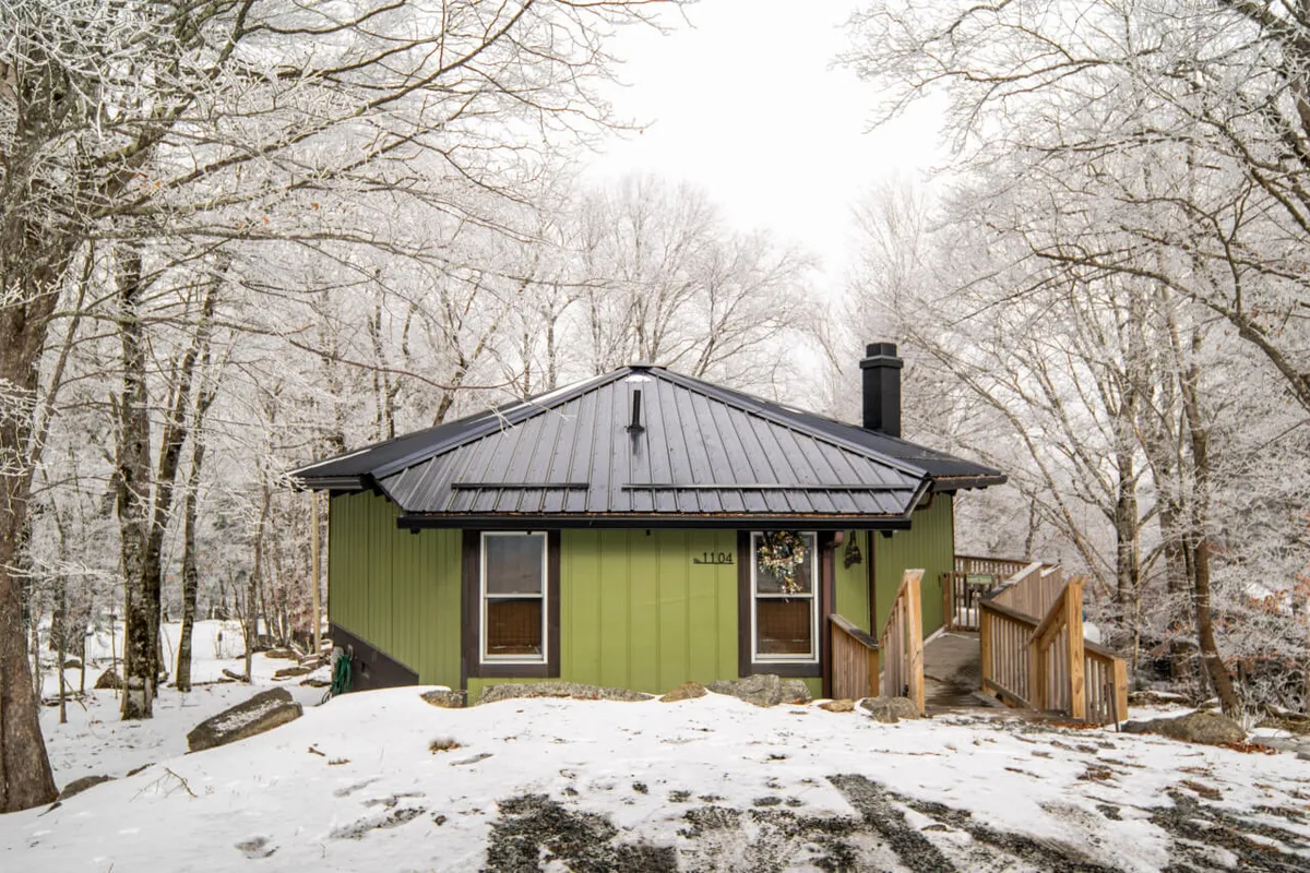 The image showcases the charming green exterior of a cozy cottage nestled among snow-covered trees on Beech Mountain. Wooden stairs lead up to a welcoming porch, inviting guests to relax with hot cocoa while enjoying the wintry landscape. The serene setting hints at outdoor fun like building snowmen and exploring nearby trails.
