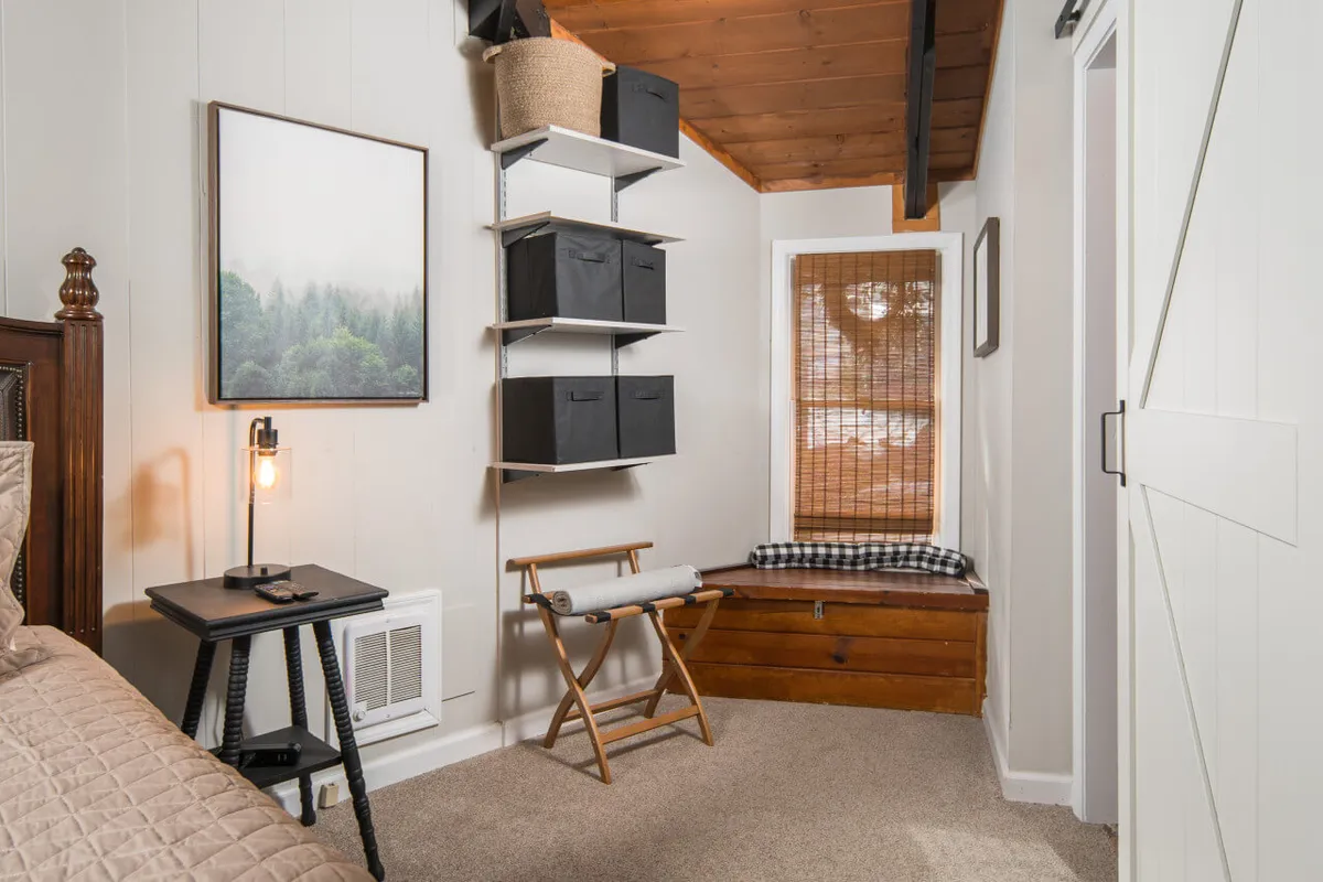 A serene bedroom with a wooden bed frame, framed landscape art, and a window seat with a cushion. The soft natural light and well-organized storage create a restful, private setting, perfect for recharging in the mountains.