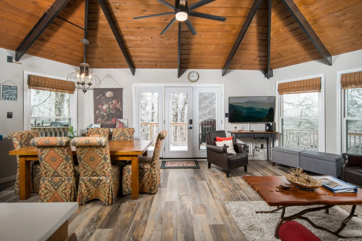 A view that ties together the dining area, living space, and kitchen. The wooden dining table sits beneath exposed beams, with large windows letting in natural light. A TV and sofa are visible, showing how these spaces are seamlessly connected for easy gathering and relaxing.