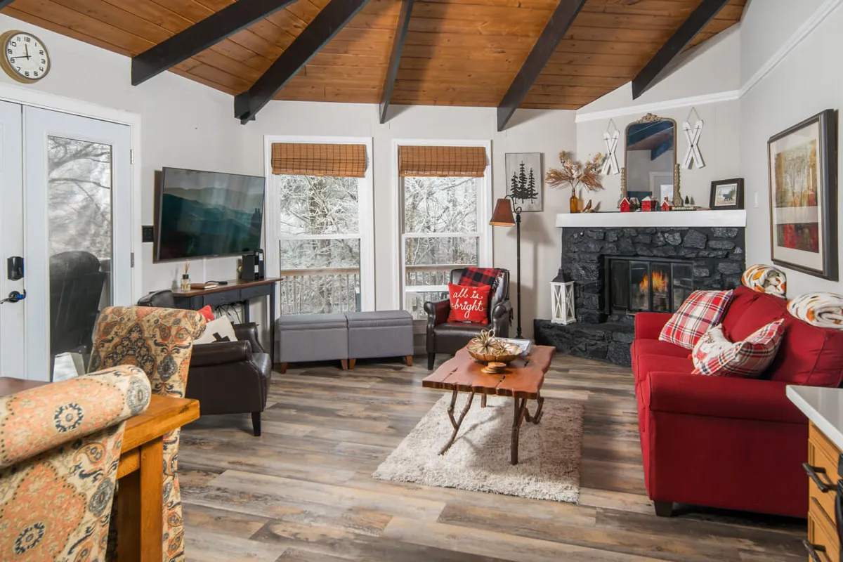 A spacious and inviting living room featuring a striking stone fireplace as the focal point. Plush red sofas are arranged for comfortable lounging, while large windows offer a snowy mountain view. Exposed wooden beams on the ceiling add rustic charm, and the overall decor is warm and modern.