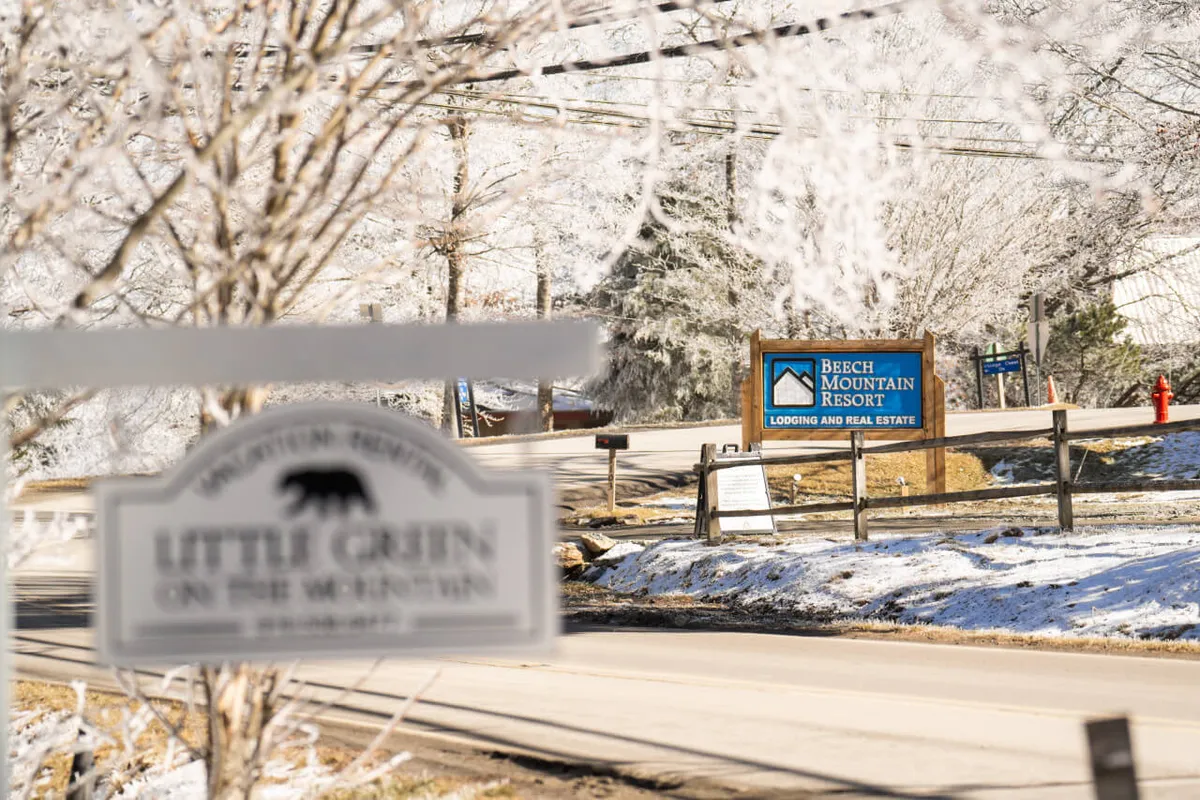 This exterior image prominently features the Beech Mountain Resort sign alongside the Little Green on the Mountain sign, set amid frost-covered trees. The winter landscape is inviting, with clear evidence of snow and the proximity to ski and adventure activities. The welcoming appearance hints at both the adventure and coziness that await guests.